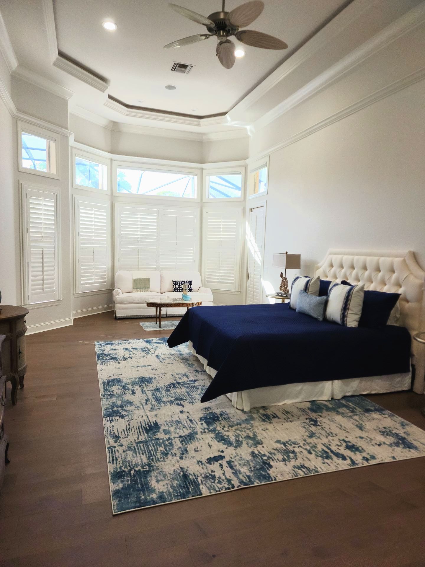 Spacious bedroom with white shutters, sofa, large blue rug, and a navy blue bedspread.