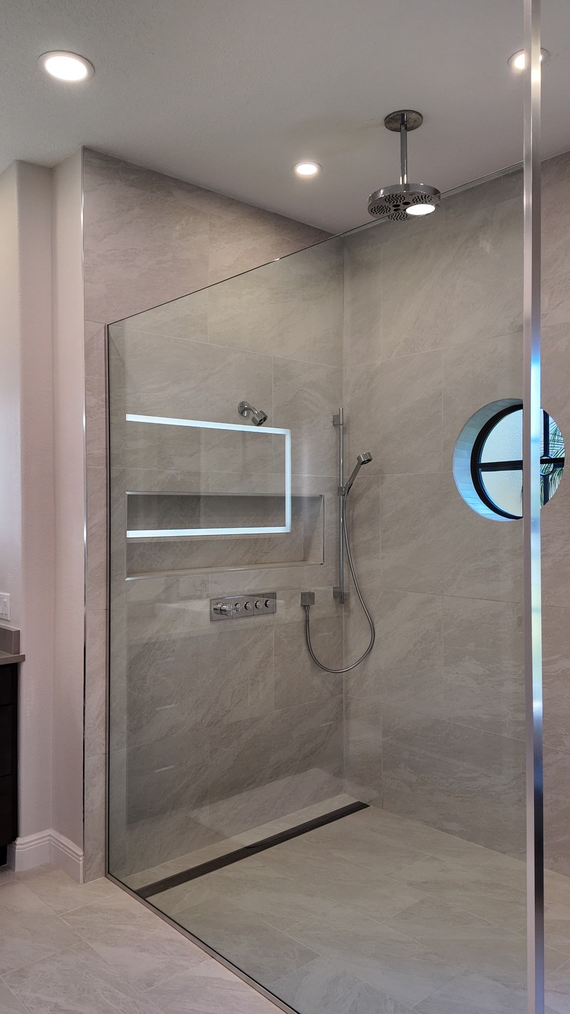 Modern gray-tiled shower with glass door, recessed shelf, and round window.